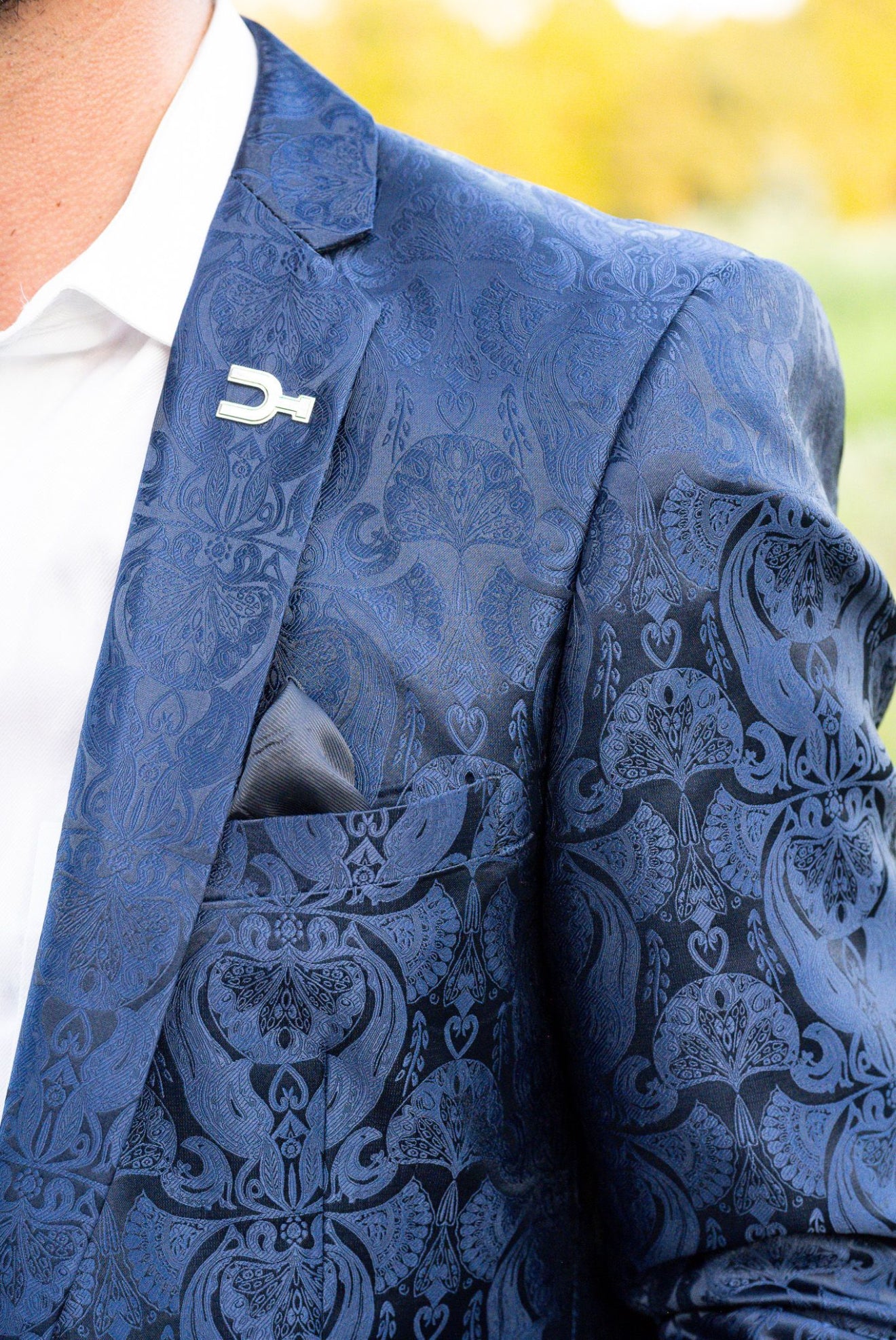 Blue patterned blazer with a white shirt and decorative lapel pin against a blurred yellow background. Cowboy sport coat with blue paisley and built in pocket square. Sterling Silver Spur lapel pin.