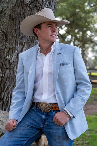 Man wearing a light blue blazer, white shirt, and cowboy hat leaning against a tree.