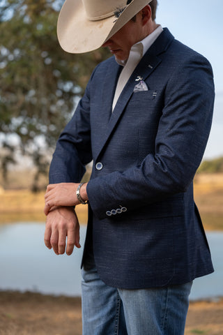 Man wearing a navy blazer with a logo, light blue jeans, and a beige cowboy hat outdoors.