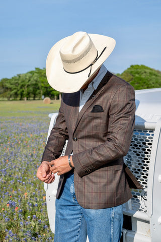 Person wearing a brown plaid jacket, blue jeans, and a white cowboy hat standing in a field with wildflowers.