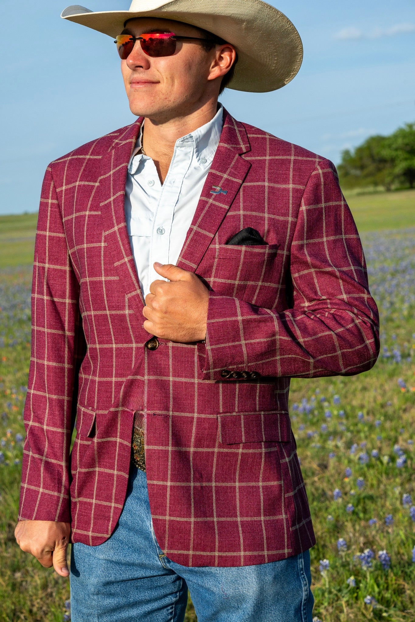 Man wearing a red plaid jacket, white shirt, and cowboy hat in a field with blue flowers.