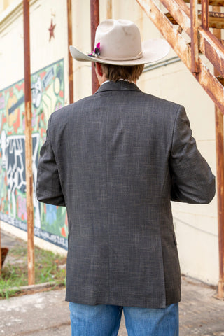 A man viewed from behind wearing a dark gray textured blazer, a cream-colored cowboy hat with a small pink flower accent, and blue jeans, standing outdoors against a rustic wall with a colorful mural and rusty metal stairs.