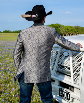 Man in a patterned jacket and cowboy hat standing next to a white truck in a field with purple flowers and green trees.