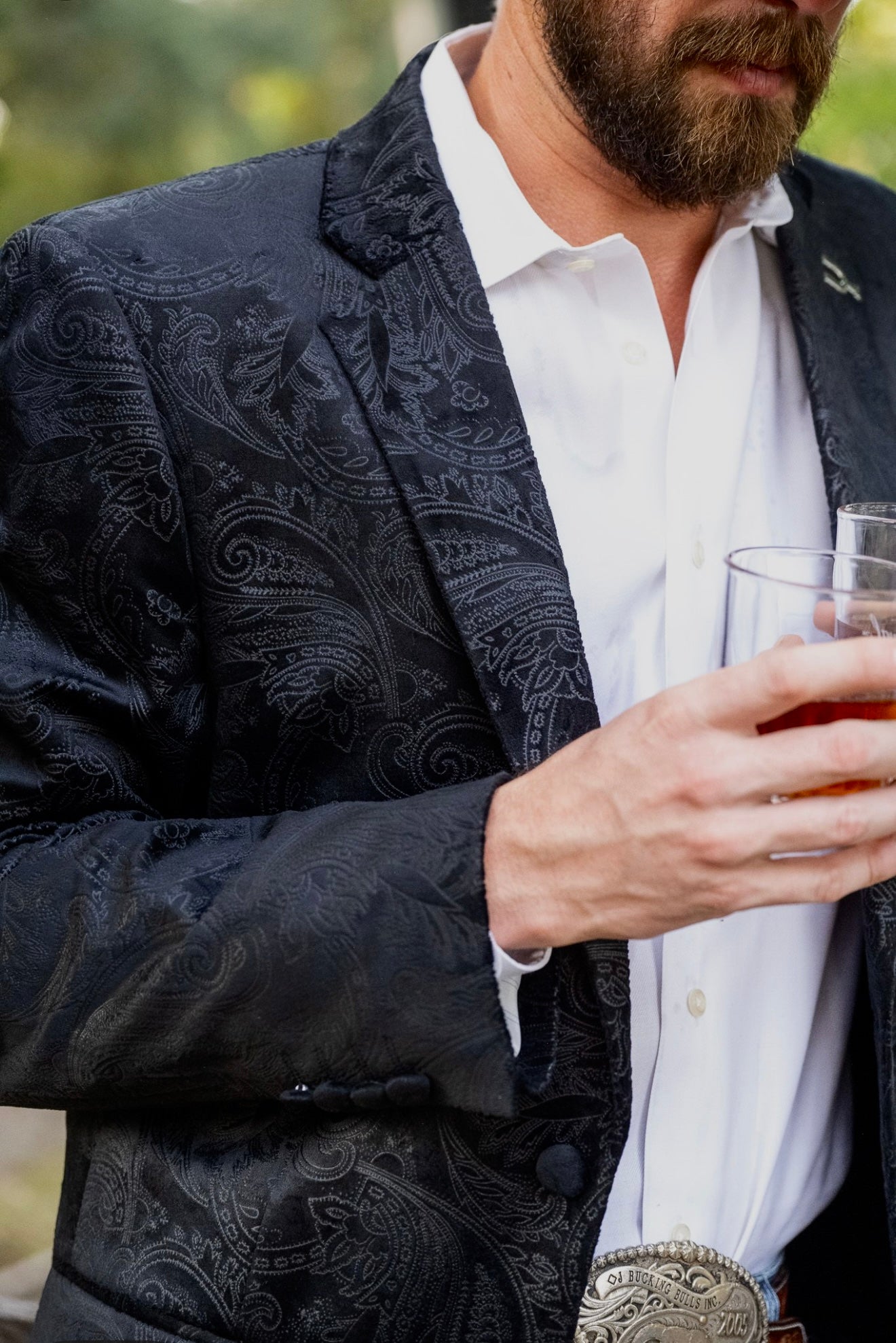 Man wearing a dark patterned blazer over a white shirt, holding a glass of amber liquid.
