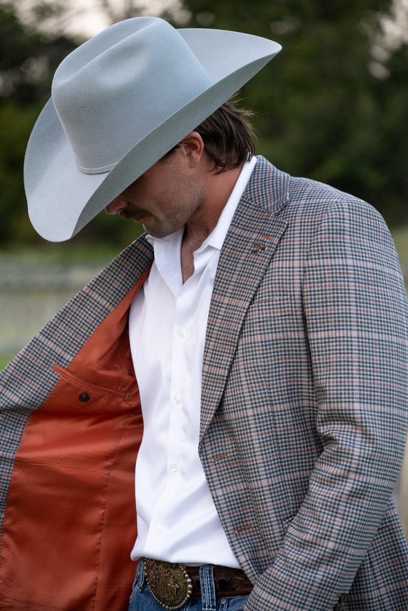 Man wearing a gray cowboy hat, plaid jacket, and white shirt outdoors.