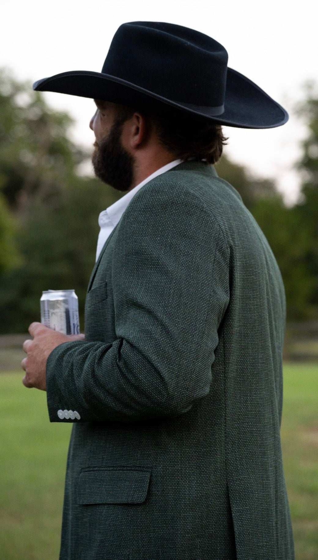 Man wearing a dark green coat and black cowboy hat holding a can, standing outdoors.