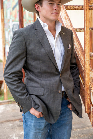 A man wearing a dark gray plaid blazer, white dress shirt, blue jeans, and a cowboy hat, standing outdoors next to a rusty metal structure.