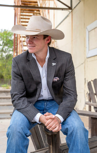 A young man wearing a light beige cowboy hat, a dark gray textured blazer, a white dress shirt, and blue jeans, sitting on a wooden porch bench.